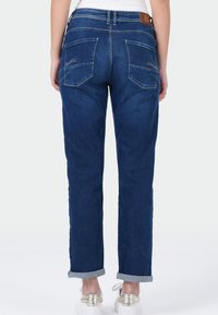 Femme portant un jean bleu foncé avec des revers et des baskets blanches avec un détail doré au talon, se tenant de dos contre un fond uni.