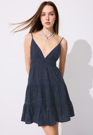 Młoda kobieta o prostych brązowych włosach, ubrana w ciemnoniebieską sukienkę bez rękawów z cienkimi ramiączkami i wijącym się haftem, stojąca na białym tle.