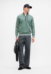 Grüner Pullover mit halbem Reißverschluss, strukturiertem Stoff und gerippten Bündchen, kombiniert mit lässigen dunklen Jeans und einer schwarzen Tasche. Eine schwarze Mütze rundet das Outfit ab.