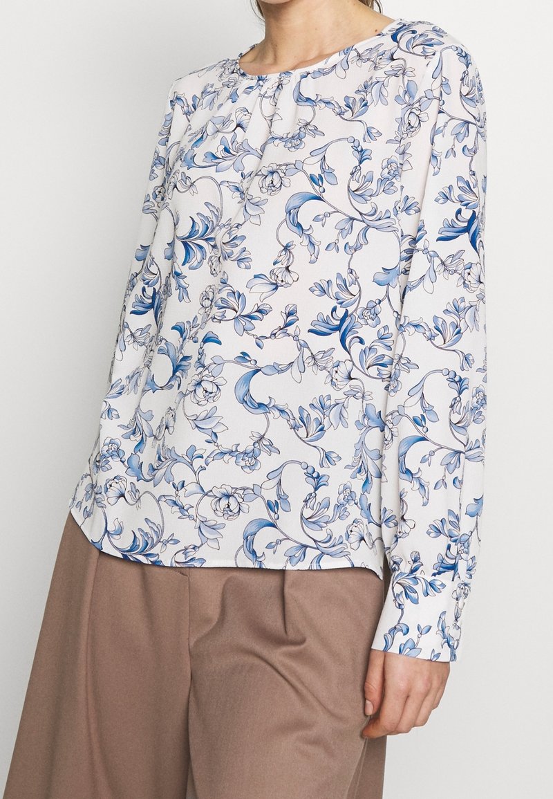 Donna con una blusa bianca a maniche lunghe con motivo floreale blu e pantaloni beige a gamba larga, in piedi contro uno sfondo semplice.