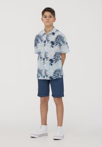 Camicia a maniche corte con bottoni di colore azzurro chiaro con motivo di foglie tropicali blu scuro, abbinata a pantaloni corti blu navy e sneakers alte bianche.