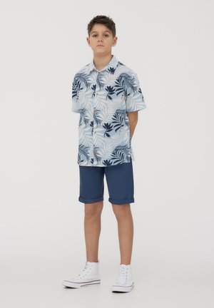 Camicia a maniche corte con bottoni di colore azzurro chiaro con motivo di foglie tropicali blu scuro, abbinata a pantaloni corti blu navy e sneakers alte bianche.