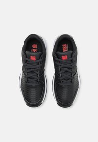 Zapatillas deportivas negras con una parte superior sintética texturizada, cordones y una suela blanca. Presentan una marca roja en la lengüeta y detalles con patrón.