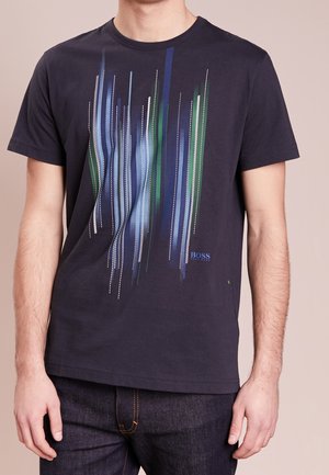 Männlicher Oberkörper trägt ein marineblaues T-Shirt mit vertikalen, abstrakten Streifen in Blau, Grün und Weiß sowie kleinem "BOSS HUGO BOSS"-Schriftzug auf der Brust.