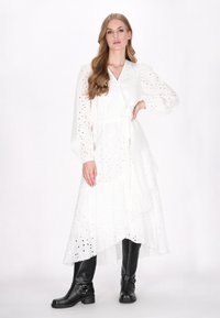 Witte jurk met lange mouwen, oogjespatroon, wikkeldesign en gelaagde rok. Gecombineerd met zwarte kniehoge laarzen met hardware-accenten.