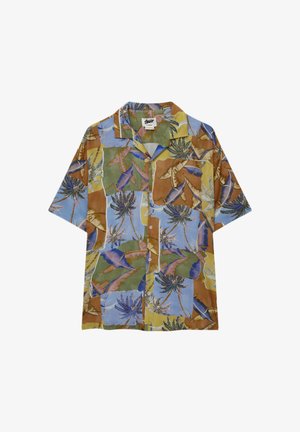 Homme portant une chemise ample à manches courtes à boutons avec un motif coloré de palmiers tropicaux, sur un top beige sans manches et un pantalon blanc.