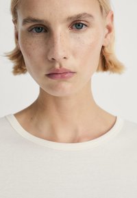 Närbild av en person med kort blont hår som bär en ljus t-shirt med rund halsringning och mjuk textur.