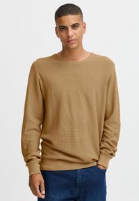 Pull en laine marron avec un motif texturé en maille gaufrée, col rond et manches à bords côtelés. Associé à un jean en denim bleu.