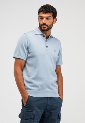 Mann mit Bart und lockigem Haar, trägt ein hellblaues kurzärmeliges Poloshirt und dunkelblaue Cargo-Shorts, Hände in den Taschen, schaut nach unten und rechts.