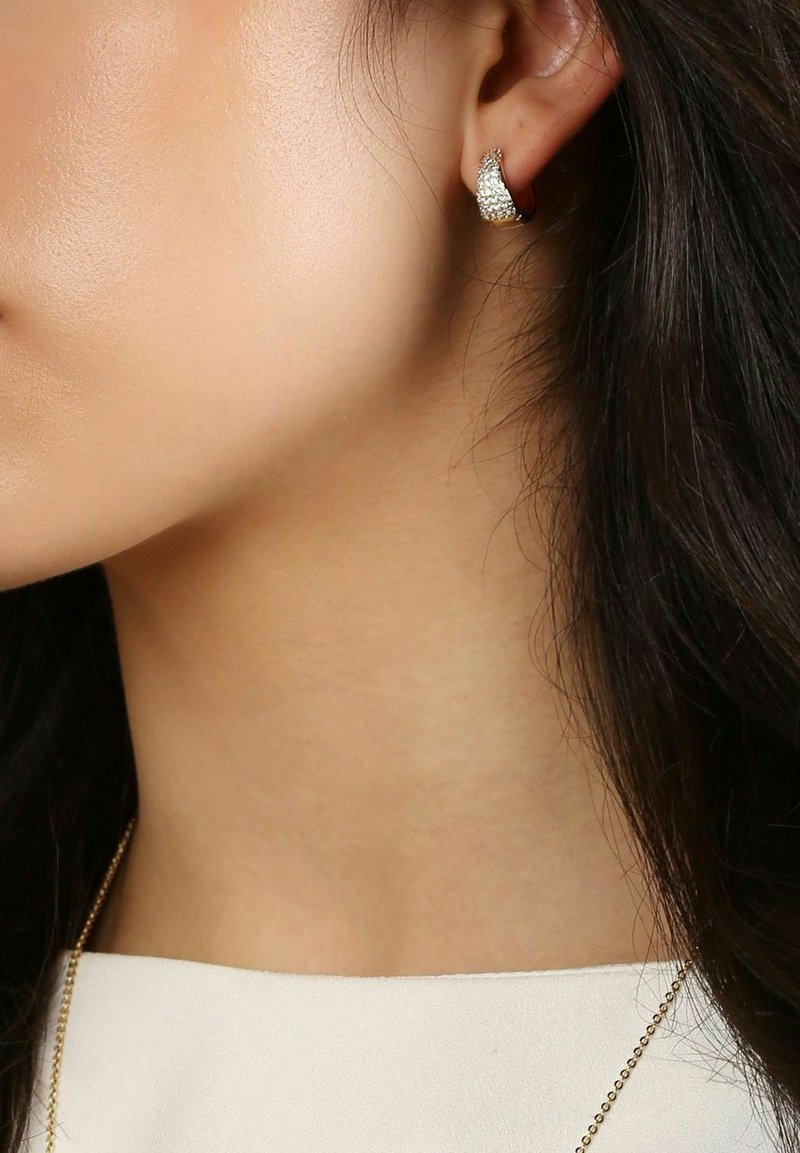 Goldfarbene Ohrringe mit geschwungenem Design, versehen mit strukturierter Oberfläche und glitzernden Akzenten, die mit einem Stift am Ohr befestigt sind.