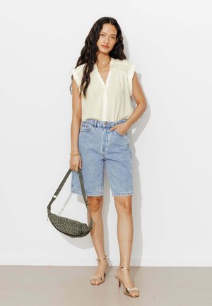 Femme portant un chemisier blanc sans manches et un short en jean longueur genou, tenant un sac à main imprimé léopard, debout contre un mur blanc.