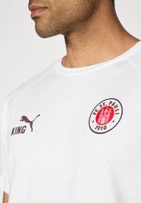 Bijela majica kratkih rukava s okruglim izrezom, koja sadrži smeđi Puma logo i natpis "KING", zajedno s kružnim oznakom FC St. Paulija.