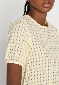 Blusa amarela às bolas com mangas curtas bufantes, apresentando um decote redondo e um tecido texturizado com quadrados em amarelo claro e castanho.
