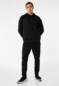 Czarna bluza z kapturem i sznurkiem, w połączeniu z czarnymi dopasowanymi spodniami. Lekki materiał, minimalistyczny design i czarne sneakersy z kontrastową podeszwą.