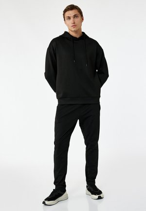 Sudadera negra con cordones, combinada con pantalones negros ajustados. Tejido ligero, diseño minimalista y zapatillas negras con suela de contraste.