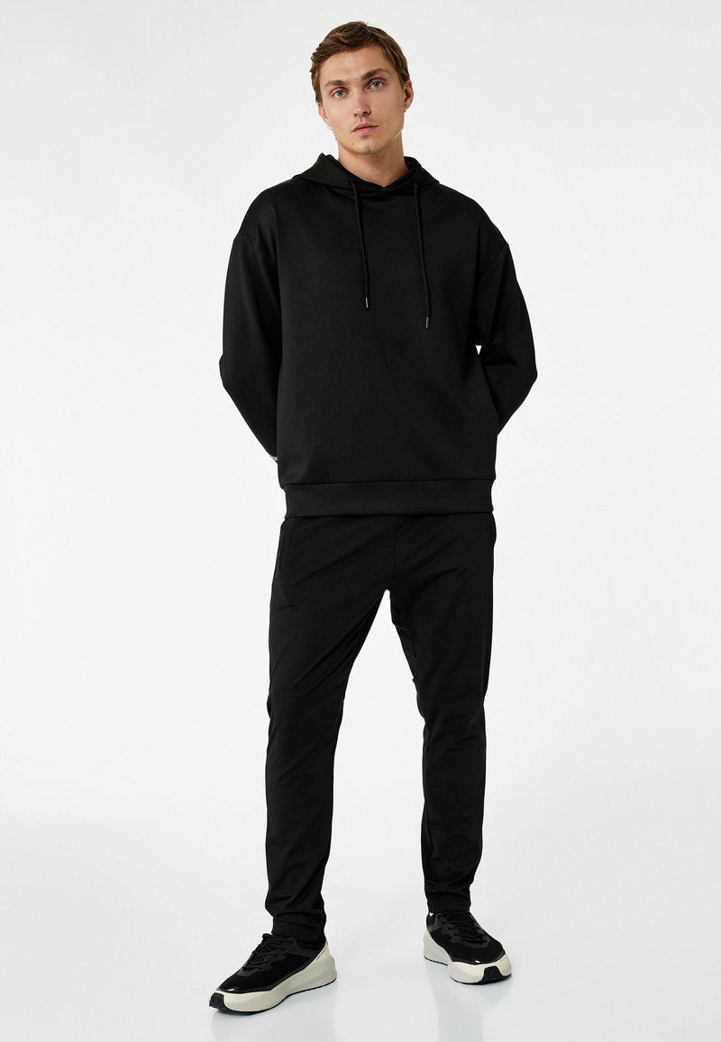 Czarna bluza z kapturem i sznurkiem, w połączeniu z czarnymi dopasowanymi spodniami. Lekki materiał, minimalistyczny design i czarne sneakersy z kontrastową podeszwą.