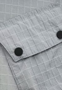 Zbliżenie jasnoszarej tkaniny z białym szwem w kratkę i klapką zapinaną na dwa czarne, okrągłe guziki.