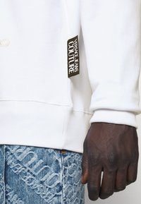 Vit sweatshirt med ribbad mudd, med en svart etikett som säger "VERSACE JEANS COUTURE" på sidan, i kombination med mönstrad ljusblå denim.