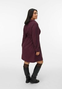 Langere mouwen, bordeaux rode corduroy jurk met een ceintuur in de taille, voorzien van een kraag en knoopdetails, gecombineerd met kniehoge zwarte laarzen.