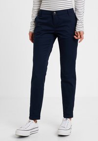 Námořnické chinos kalhoty s rovnými nohavicemi, vybavené předním zapínáním na knoflík, poutky na opasek a hladkým bavlněným povrchem, v kombinaci s bílými teniskami.