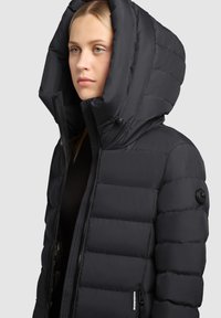 Black puffer jacket with a large hood, quilted texture, zippered front, and a logo patch on the left sleeve. Smooth, insulated material.