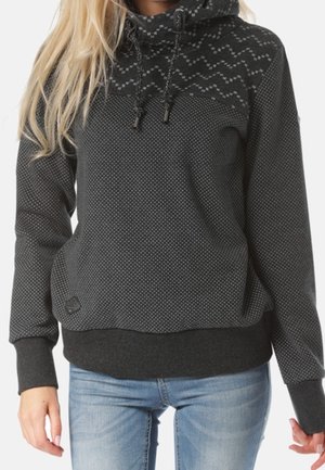 Vrouw draagt een donkergrijze pullover met capuchon met kleine lichtgrijze stippen en een zigzagpatroon op het schouderstuk, gecombineerd met lichtblauwe spijkerbroek.