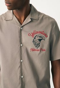 Chemise à manches courtes gris clair avec un devant boutonné. Présente une broderie rouge "Yellowstone" et un dessin d'aigle noir.