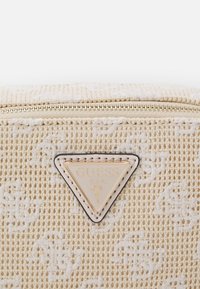 Béžová tkaná kabelka s texturovaným vzorem, zlatou trojúhelníkovou nášivkou a hladkým zipem přes horní okraj.