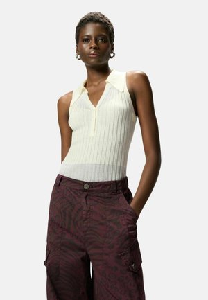 Donna che indossa una canotta crema a coste senza maniche con colletto e pantaloni cargo bordeaux a motivo, in piedi con una mano in tasca.