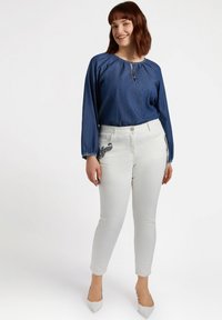 Blusa de mezclilla con escote fruncido, pantalones blancos ajustados con bordado negro en los bolsillos y tacones blancos de punta.