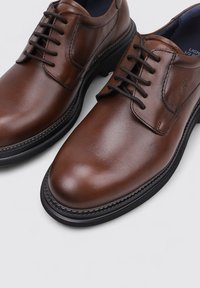 Zapatos de cuero marrón con acabado suave, puntera redonda, diseño de cordones y suela de goma negra con detalle de costura a lo largo del borde.