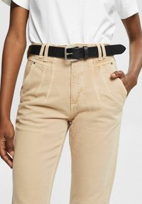 Pantalons beiges en tissu texturé, avec des plis à l'avant, des poches latérales et une ceinture en cuir noir avec une boucle en argent.