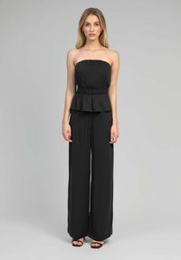 Femme aux longs cheveux blonds portant un top noir sans bretelles et un pantalon noir ample, se tenant contre un fond gris.