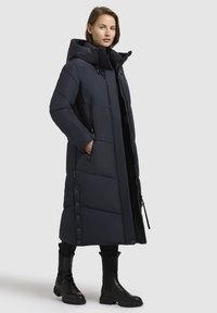 Navy blue puffer coat with a hood, featuring a long silhouette, zippered pockets, and snap-button accents along the side.