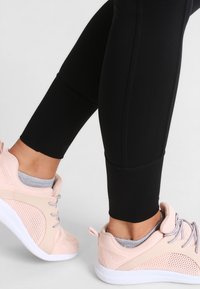 Lichtroze sneakers met mesh accenten, ronde neus en witte zool, gecombineerd met zwarte legging; grijze sokken zichtbaar boven de schoen.