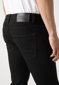 Jeans de mezclilla negra con un parche de cuero con marca en relieve, bolsillos traseros inclinados y un corte ajustado. Textura lisa y detalles mínimos.