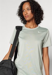 Hele roheline Nike lühikeste varrukatega särk sünteetilisest kangast, millel on ümmargune kaelus ja valge logo. Must kott on õlal rippumas.