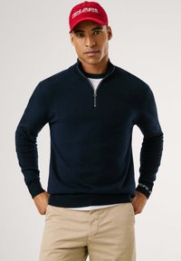 Felpa blu navy con zip in morbido tessuto a maglia, con polsini e orlo a coste, indossata su una camicia bianca, abbinata a pantaloni beige. Un cappellino rosso completa l'outfit.
