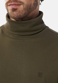 Olivgrüner Rollkragenpullover mit glatter Textur, der ein dezentes Logo-Detail auf der Brust aufweist und mit einer enganliegenden Silhouette gestaltet ist.