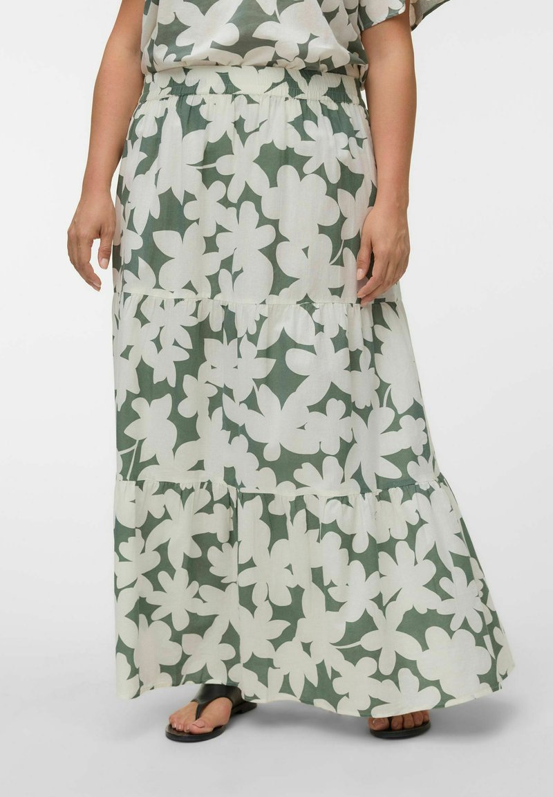 Jupe longue verte à volants avec grand imprimé floral blanc portée avec des sandales noires sur une personne debout.