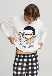 Camicia bianca a maniche lunghe con una grafica di un bradipo in blu e marrone, con la scritta "ANCHE CINQUE MINUTI..." Pantaloni del pigiama a scacchi bianco e nero.