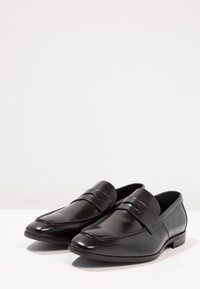 Mocassins pour hommes en cuir noir avec une finition élégante et polie, ornés d'une sangle décorative sur le dessus, présentés sur une surface blanche.