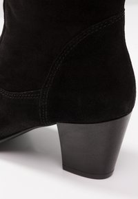 Bottine en suede noir avec un talon bloc empilé et des coutures visibles sur une surface lisse, contre un fond blanc.