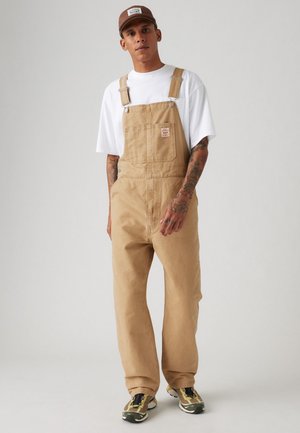 Salopette en coton beige avec des bretelles ajustables et une poche avant, associée à un T-shirt blanc et des baskets avec des accents verts.