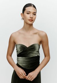 Junge Frau mit nach hinten gekämmtem Haar, die goldene Creolen und ein grünes Samt-Top ohne Träger trägt, mit den Händen auf der Taille vor einem schlichten Hintergrund.