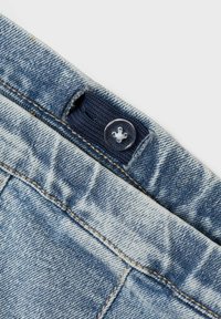 Denim tkanina v svetlo modri barvi, prikazuje bližnji posnetek zapiranja na gumb z mornarsko modrim poudarkom. Vidni so detajli šivanja in teksture.