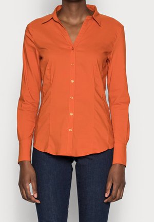 Persoon die een nauwsluitend oranje overhemd met lange mouwen en kraag draagt, gecombineerd met donkerblauwe jeans, staand tegen een effen achtergrond.