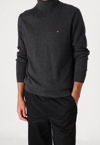 Mann in einem dunkelgrauen Rollkragenpullover mit einem kleinen rot-weiß-blauen Logo auf der Brust und schwarzen Hosen, Hände entspannt an den Seiten.
