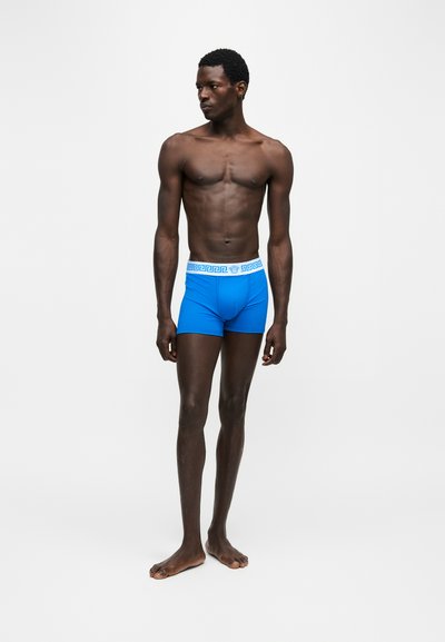Homme grand à la peau mate portant un boxer bleu vif avec une ceinture blanche ornée d'un motif de clé grecque, debout pieds nus devant un fond uni.