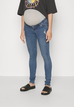 Jeans skinny de maternidade em denim azul médio, com um ligeiro desvanecimento e um remendo desgastado. Combinados com uma t-shirt gráfica solta preta e sandálias pretas.